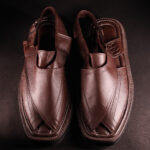Top view of a pair of handcrafted brown leather Peshawari chappals featuring a traditional cross-strap design and detailed stitching, placed on a dark textured background.