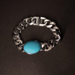 A silver chain bracelet with a bold, oval turquoise stone centrepiece, displayed on a dark textured background.