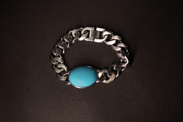 A silver chain bracelet with a bold, oval turquoise stone centrepiece, displayed on a dark textured background.
