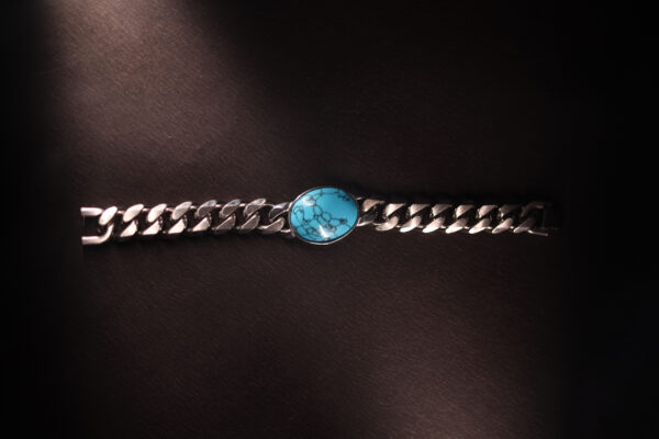 Flat-lay image of a silver Cuban link bracelet with a marbled turquoise stone centrepiece, placed on a dark textured background with dramatic lighting.