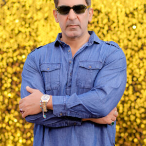 A man wearing a denim Pathani suit, styled with sunglasses and accessories, standing confidently against a vibrant golden sequin background.