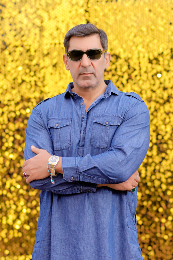A man wearing a denim Pathani suit, styled with sunglasses and accessories, standing confidently against a vibrant golden sequin background.